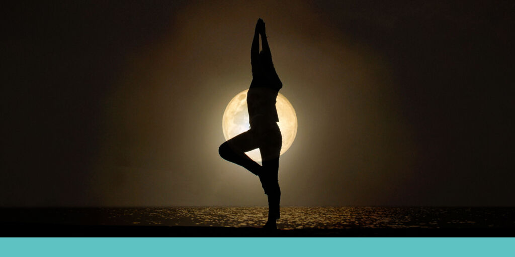A woman in a yoga pose on the beach with the moon in the background.